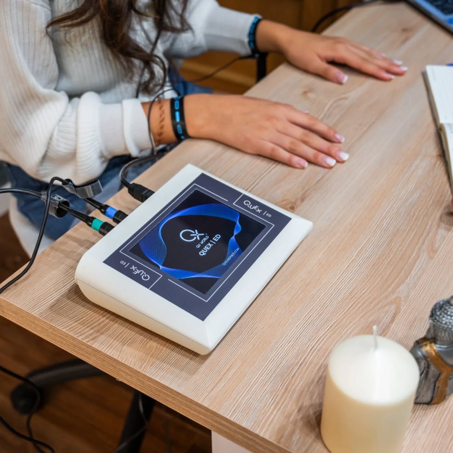 Una mujer en un escritorio utiliza un dispositivo con una pantalla, cables y una vela cerca.