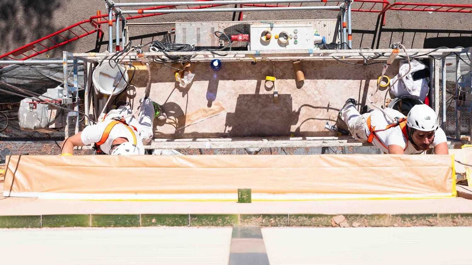 Dos trabajadores con arneses y cascos blancos están de pie sobre una plataforma de andamio suspendida contra una pared.
