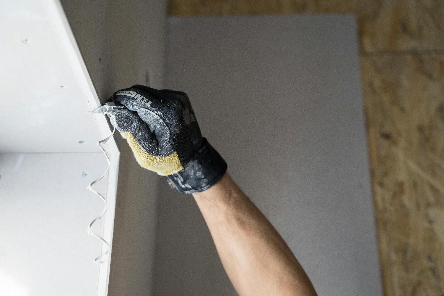 Un trabajador, con guantes de trabajo, corta un trozo de placa de yeso blanca en una obra de construcción interior.