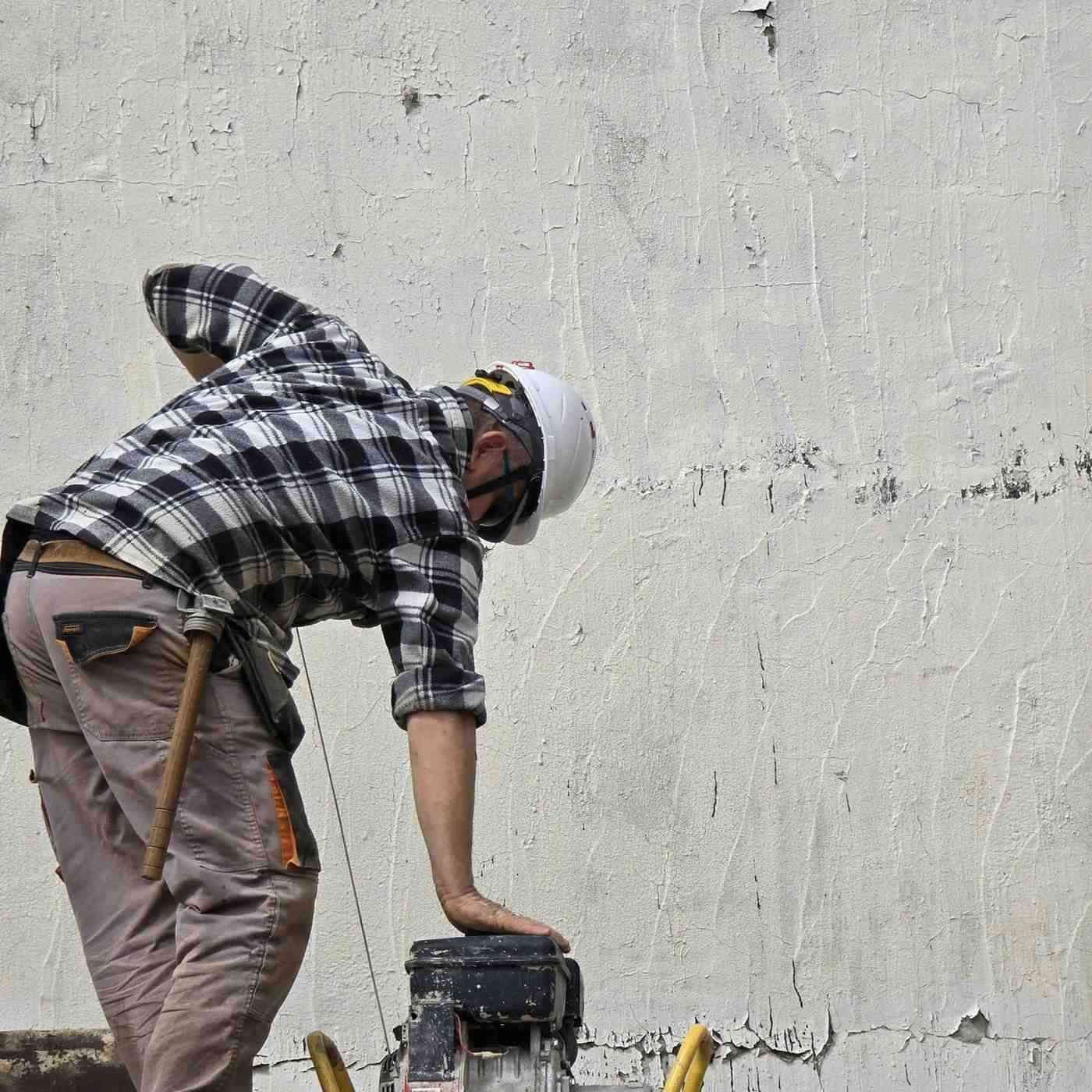 Un trabajador con camisa de cuadros y casco se inclina, manejando una máquina de construcción contra una pared desgastada.