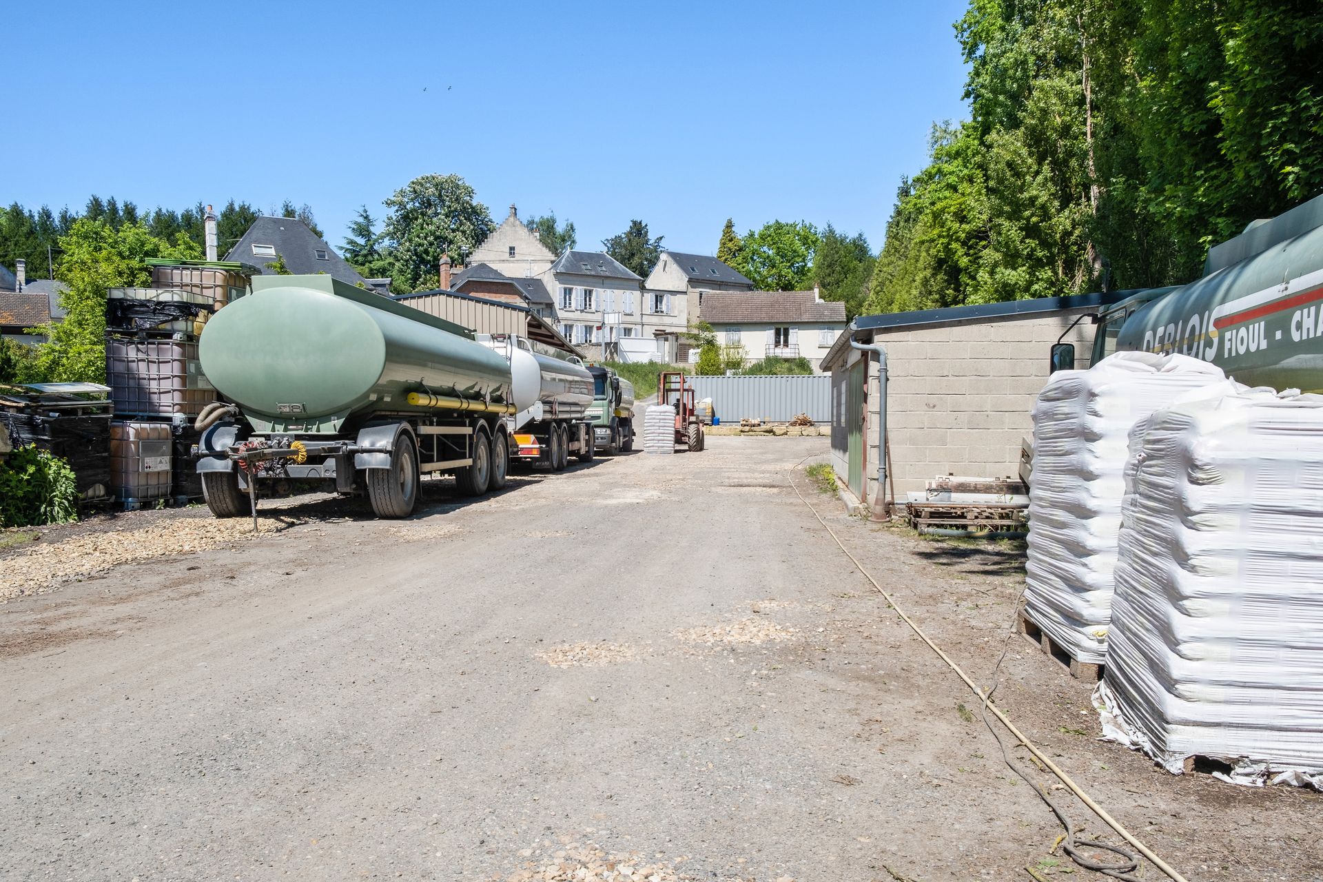 Un terrain vague avec de gros camions-citernes garés à gauche et une pile de sacs de matériaux à droite, près d'un bâtiment.