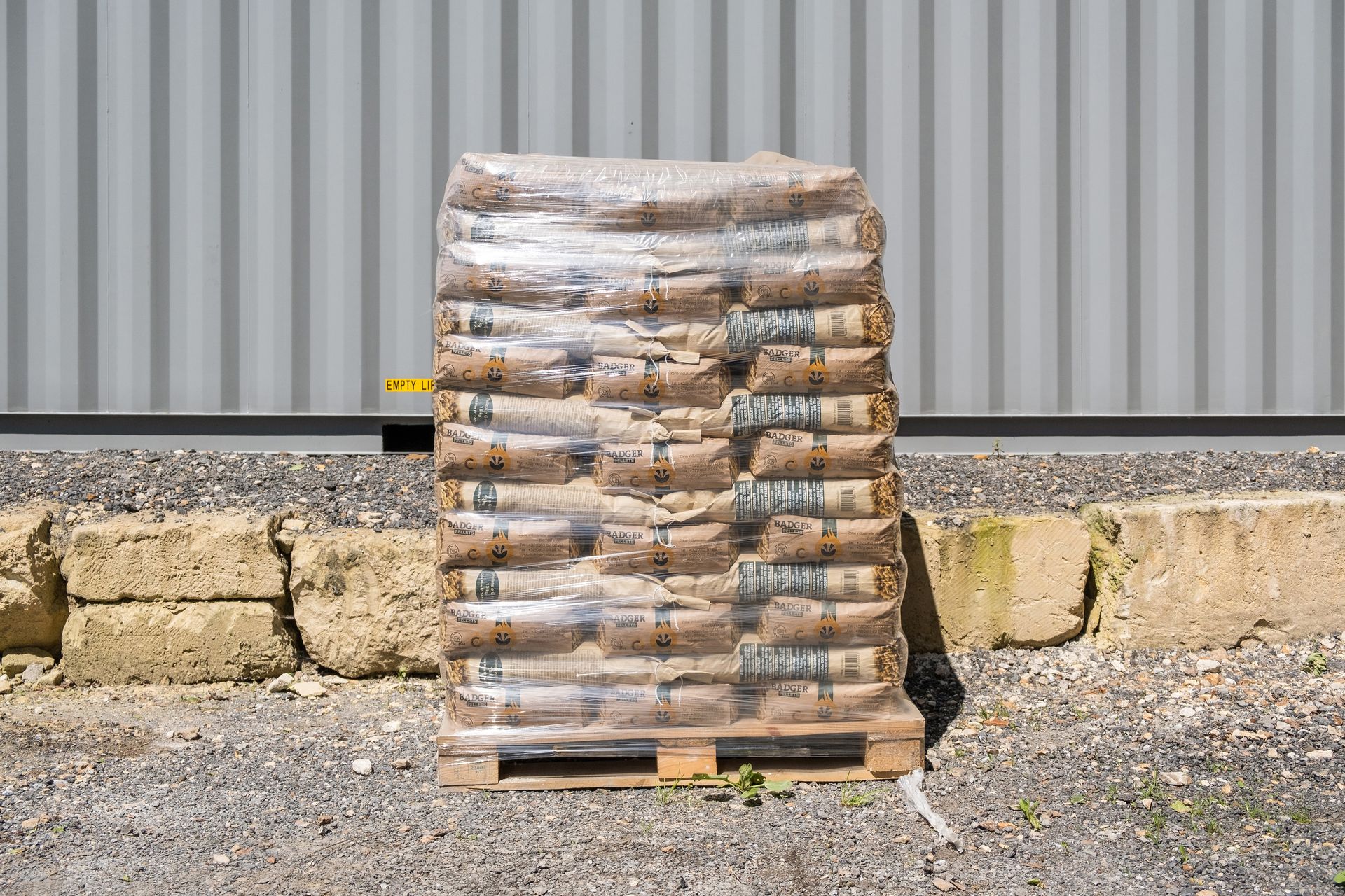Une palette de sacs bruns empilés et filmés, adossée à un mur de tôle ondulée et à des blocs de pierre.