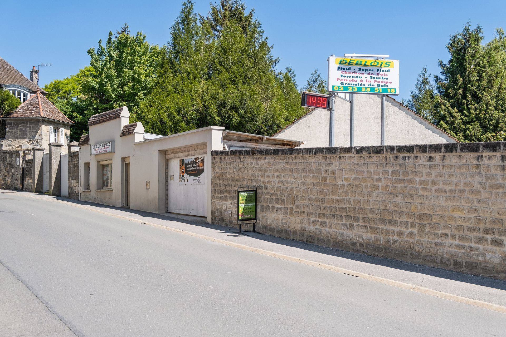Vue en bord de route d'un bâtiment en pierre avec un garage, arborant un panneau blanc au sommet d'un mur de pierre et un panneau vert devant.
