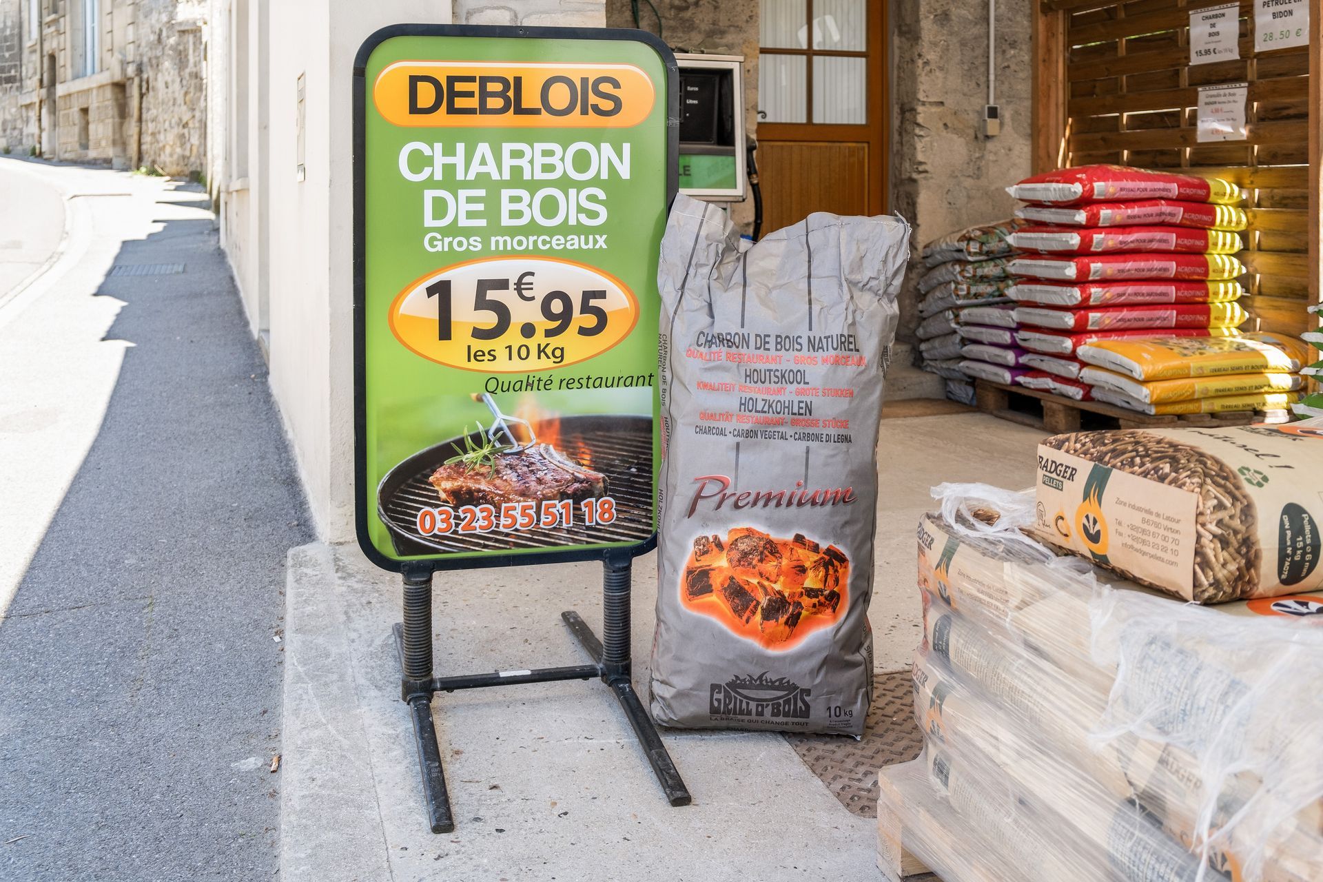 Une affiche publicitaire pour le charbon de bois Deblois (15,95 € les 10 kg) se dresse à côté d'un grand sac de charbon de bois devant un magasin.