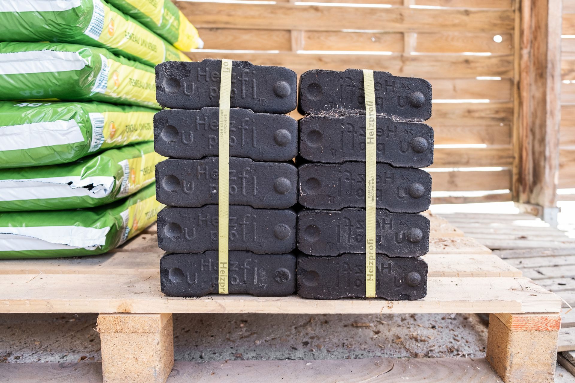 Deux piles de briquettes de combustible rectangulaires et sombres, maintenues ensemble par des sangles en plastique jaune sur une palette en bois.