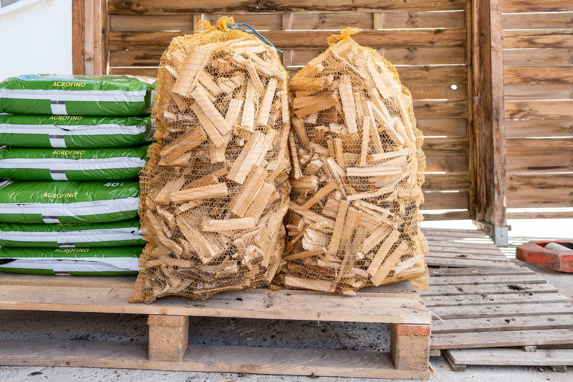 Deux sacs en filet remplis de bois de chauffage coupé sont posés sur une palette en bois à côté d'une pile de sacs en plastique verts.
