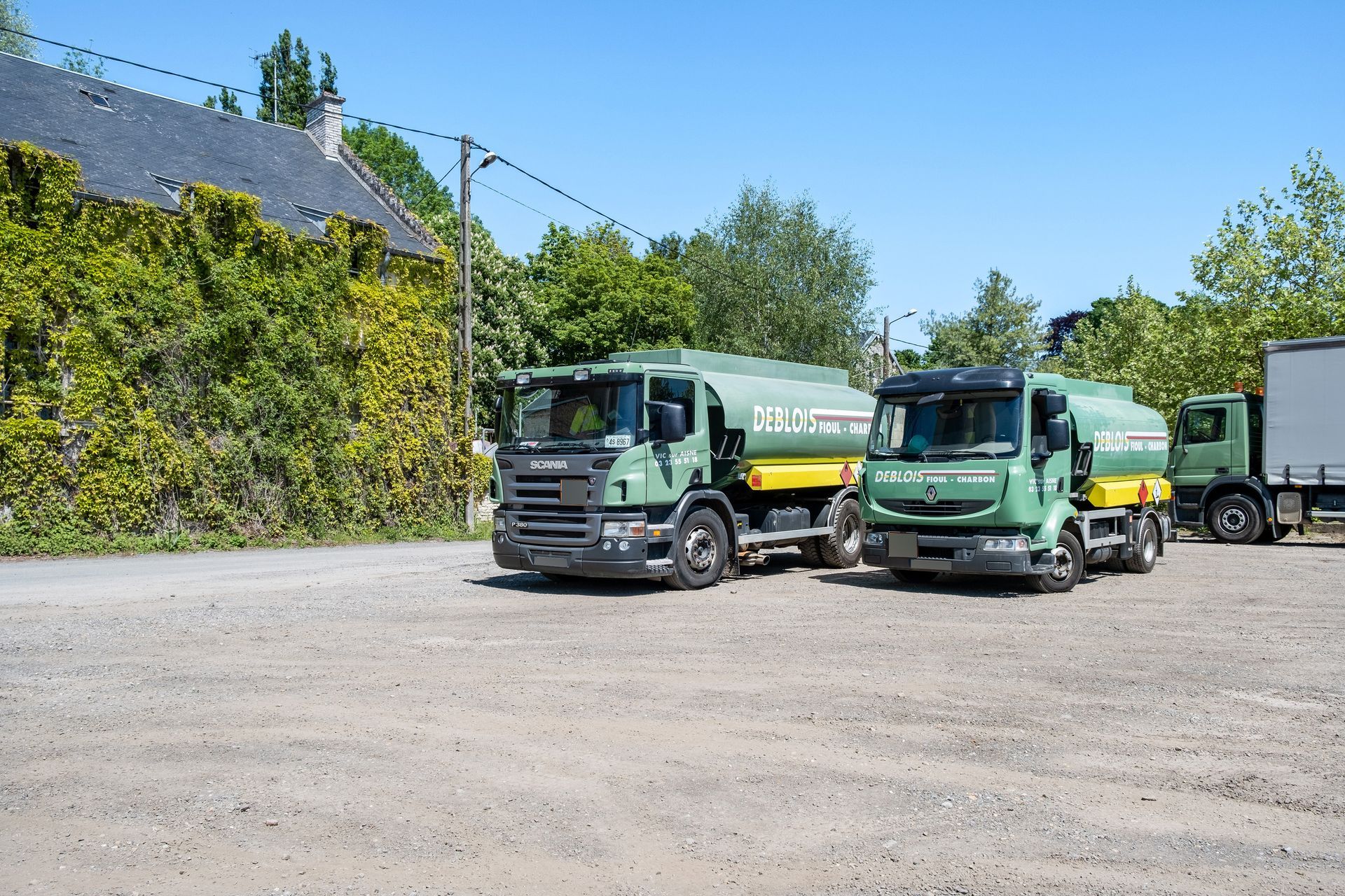 Trois camions-citernes verts et jaunes sont stationnés sur un terrain gravillonné à côté d'un bâtiment vert et d'arbres.