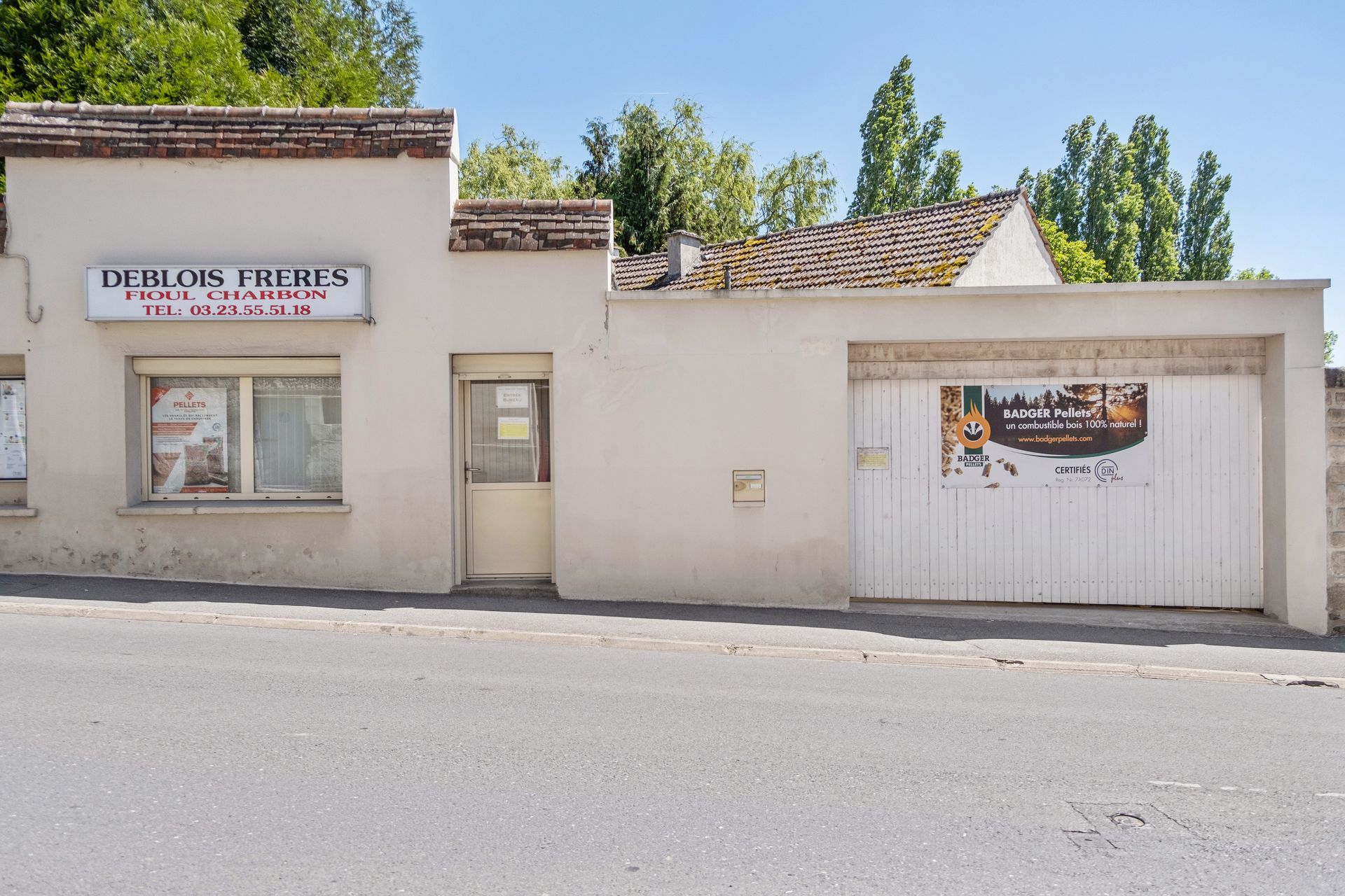 Un bâtiment beige de plain-pied avec une enseigne « Deblois Frères » et une porte de garage blanche, donnant sur une rue pavée.