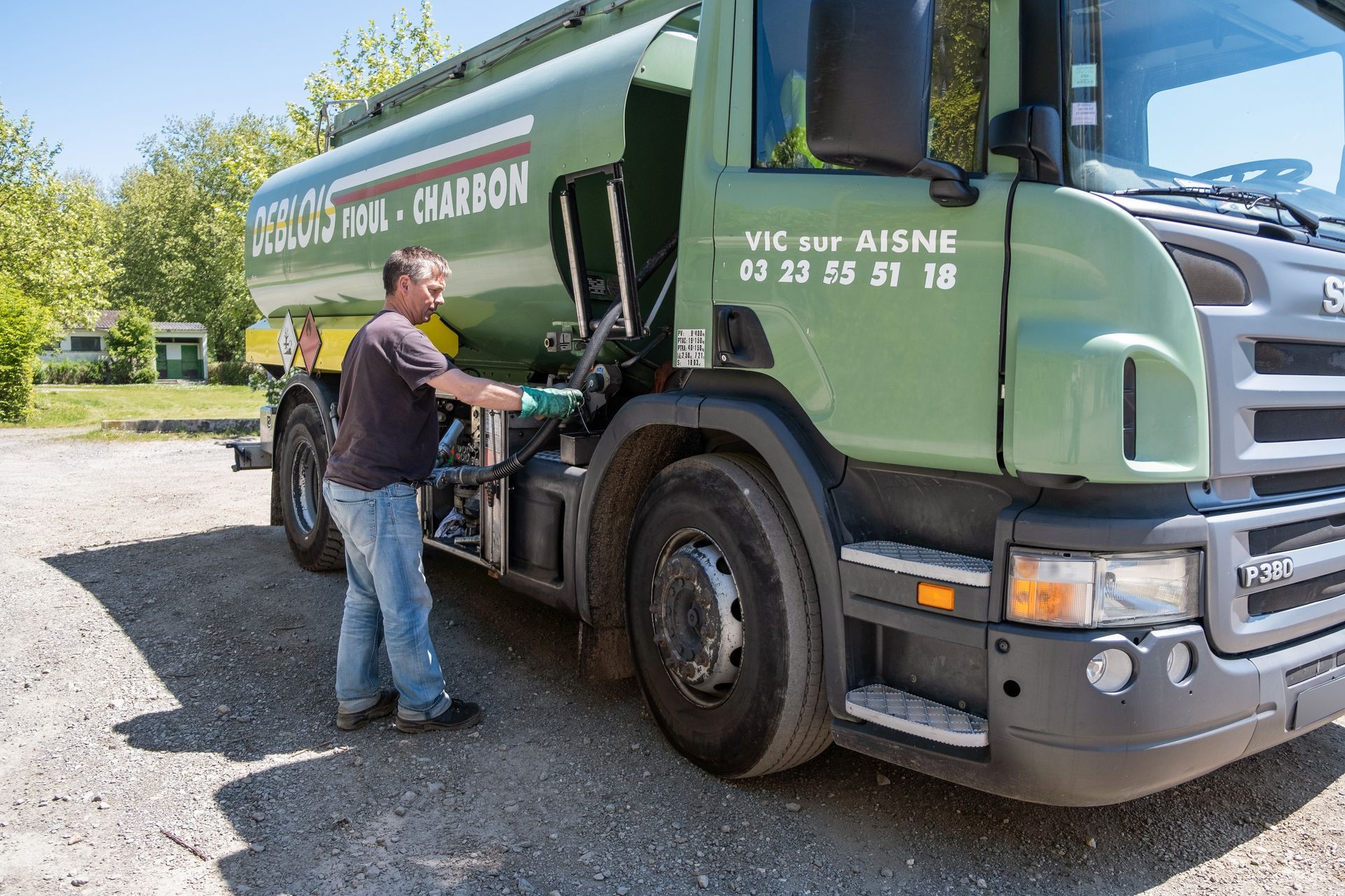 Une personne en tenue de travail actionne l'équipement d'un camion de livraison de carburant vert stationné sur un terrain gravillonné.