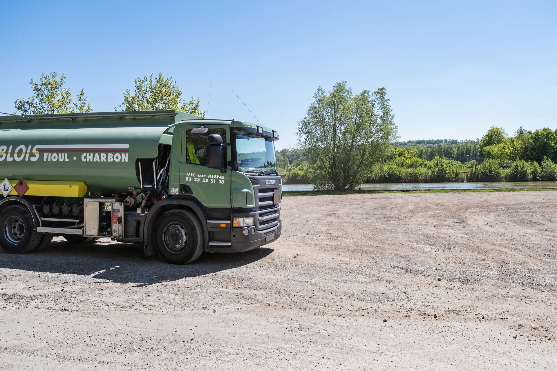 Un camion-citerne vert clair est stationné sur un terrain gravillonné près d'une rivière et d'arbres, sous un ciel bleu dégagé.