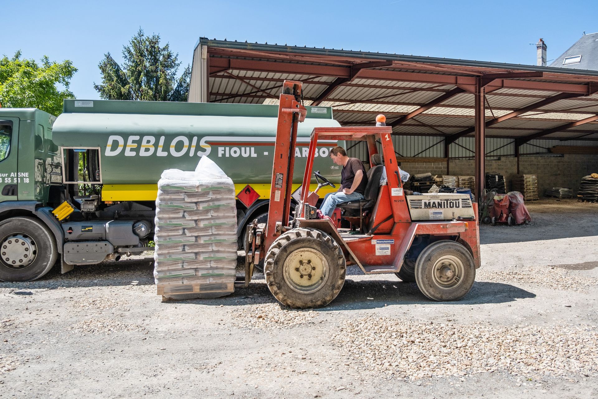 Un chariot élévateur rouge transporte une palette de matériaux ensachés devant un camion-citerne vert stationné sous un hangar industriel ouvert sur les côtés.
