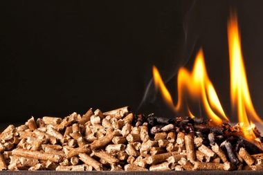 Un tas de granulés de bois, dont le côté droit prend feu et produit des flammes et de la fumée sur un fond sombre.