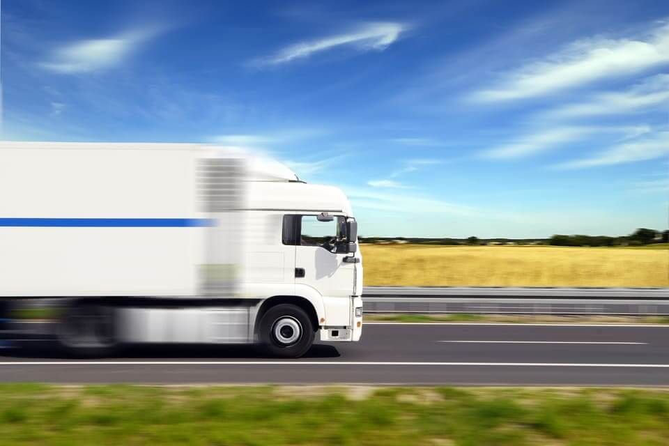 Un camion blanc roulant à vive allure sur une autoroute, avec un ciel bleu et un champ doré en arrière-plan.
