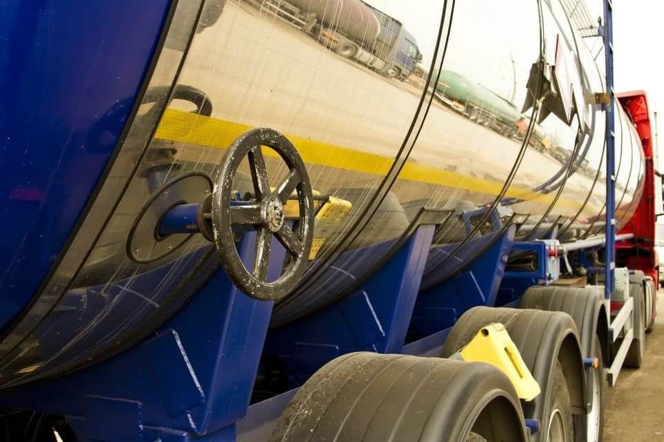 Vue latérale d'une grande semi-remorque citerne industrielle métallique brillante, avec des supports bleus et des roues noires.