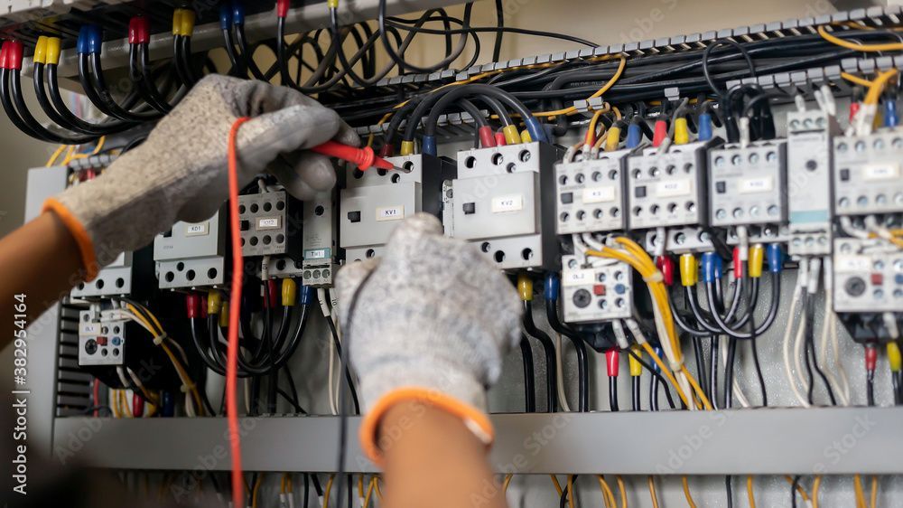 Personne testant un tableau électrique avec un multimètre, portant des gants de travail.
