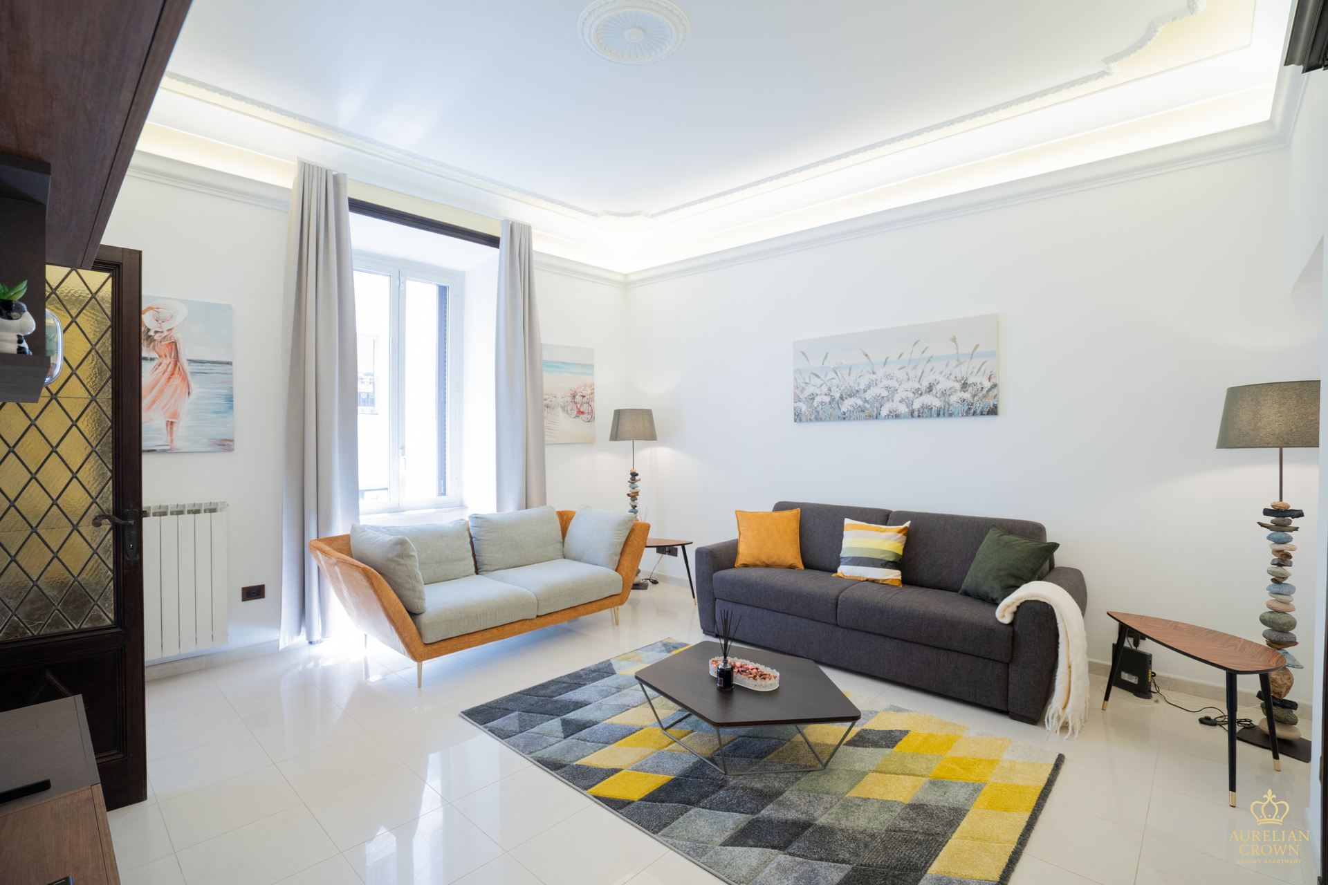 A bright living room with white walls, two sofas, and a geometric rug. Window with light gray curtains, and various decor.