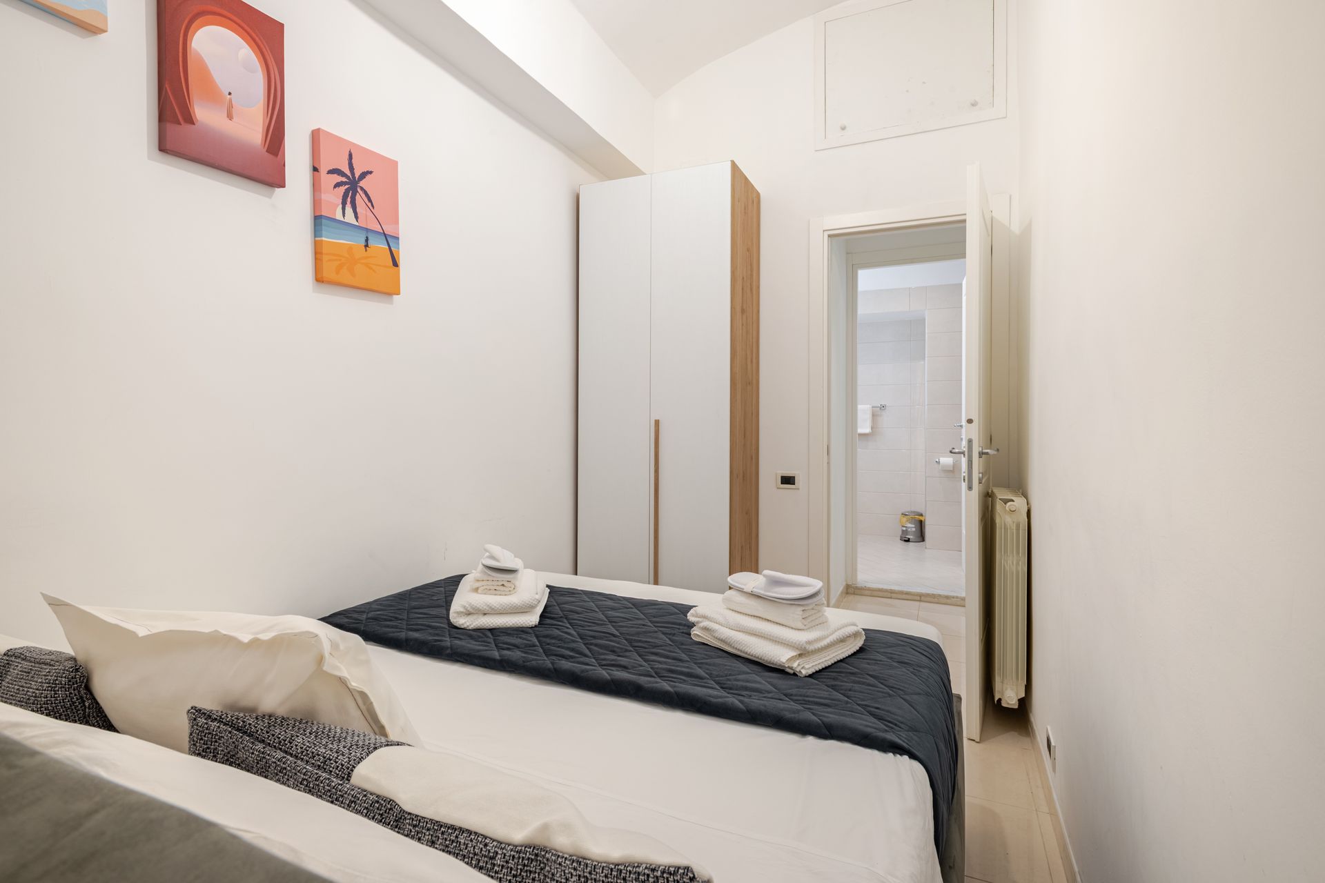 A small bedroom with a bed, closet, and open doorway to a bathroom. Artwork hangs on the white wall.