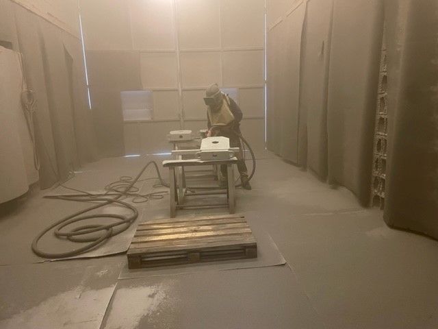 Homme en tenue de sablage dans l'atelier réalisant le sablage d'une pièce 