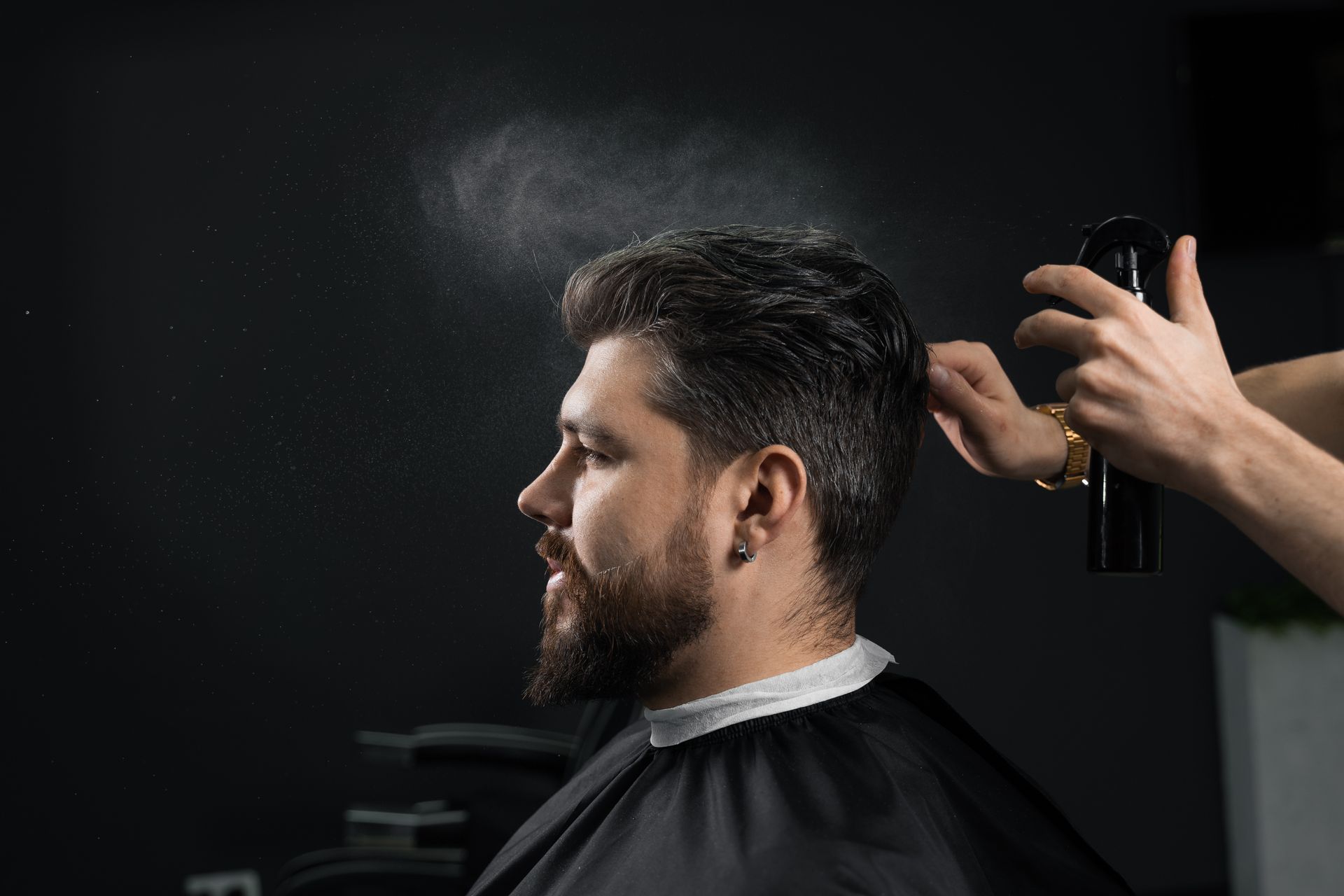 Mann im Friseurstuhl lässt sich die Haare mit Haarspray stylen. Dunkler Hintergrund, Profilansicht.