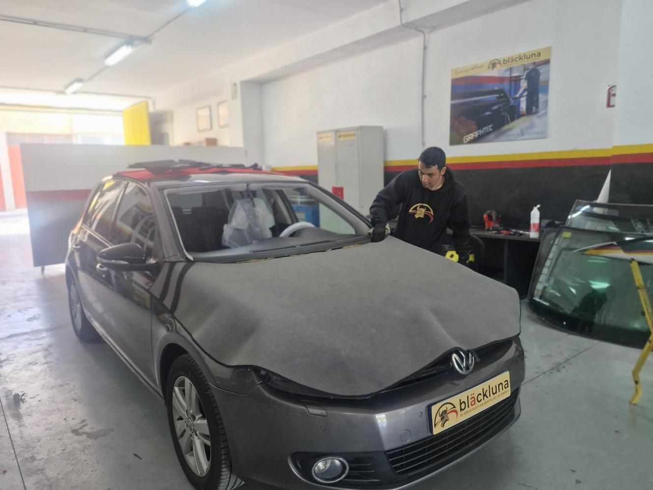 Una persona trabaja en un coche gris dentro de un garaje. El coche está cubierto con una funda protectora.