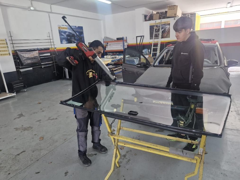 Dos personas instalan un parabrisas en un taller. Una aplica sellador, la otra observa.