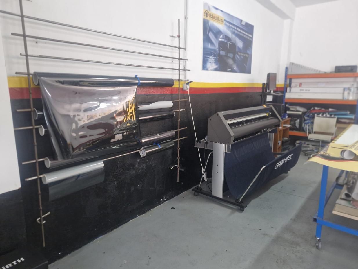 Escena interior de tienda con una impresora de gran formato, rollos de material y un estante de almacenamiento.