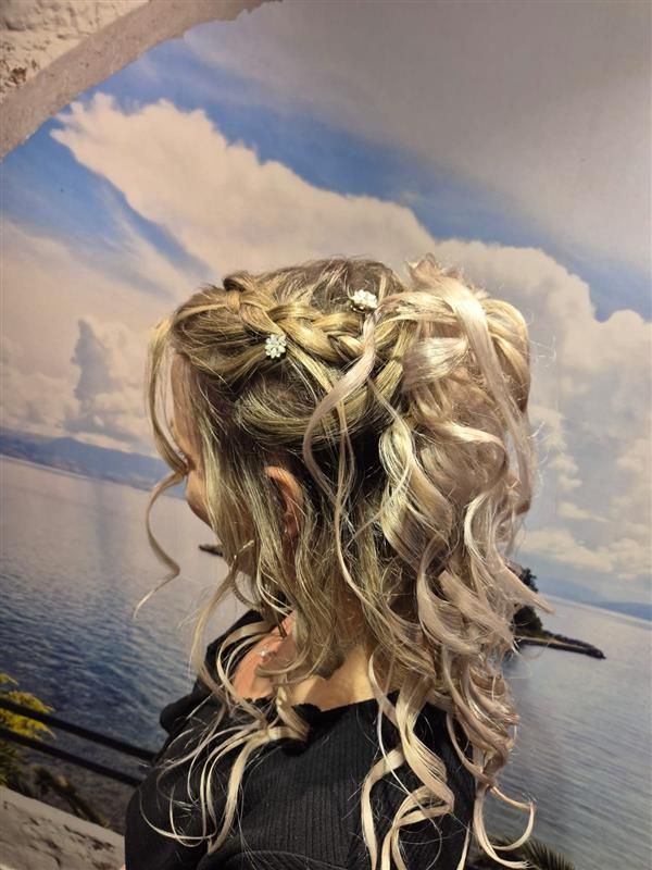 Lockige blonde Hochsteckfrisur mit geflochtenen Akzenten vor einem gemalten Himmel- und Meereswandbild