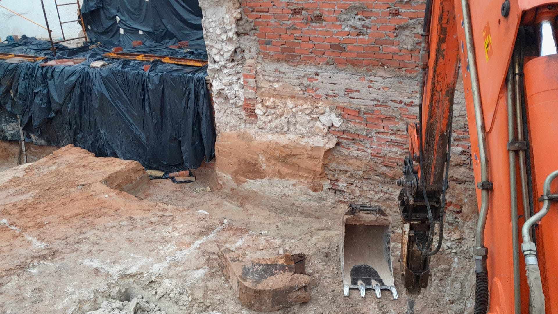 Una gran excavadora naranja está cavando un hoyo en el suelo frente a una pared de ladrillos.