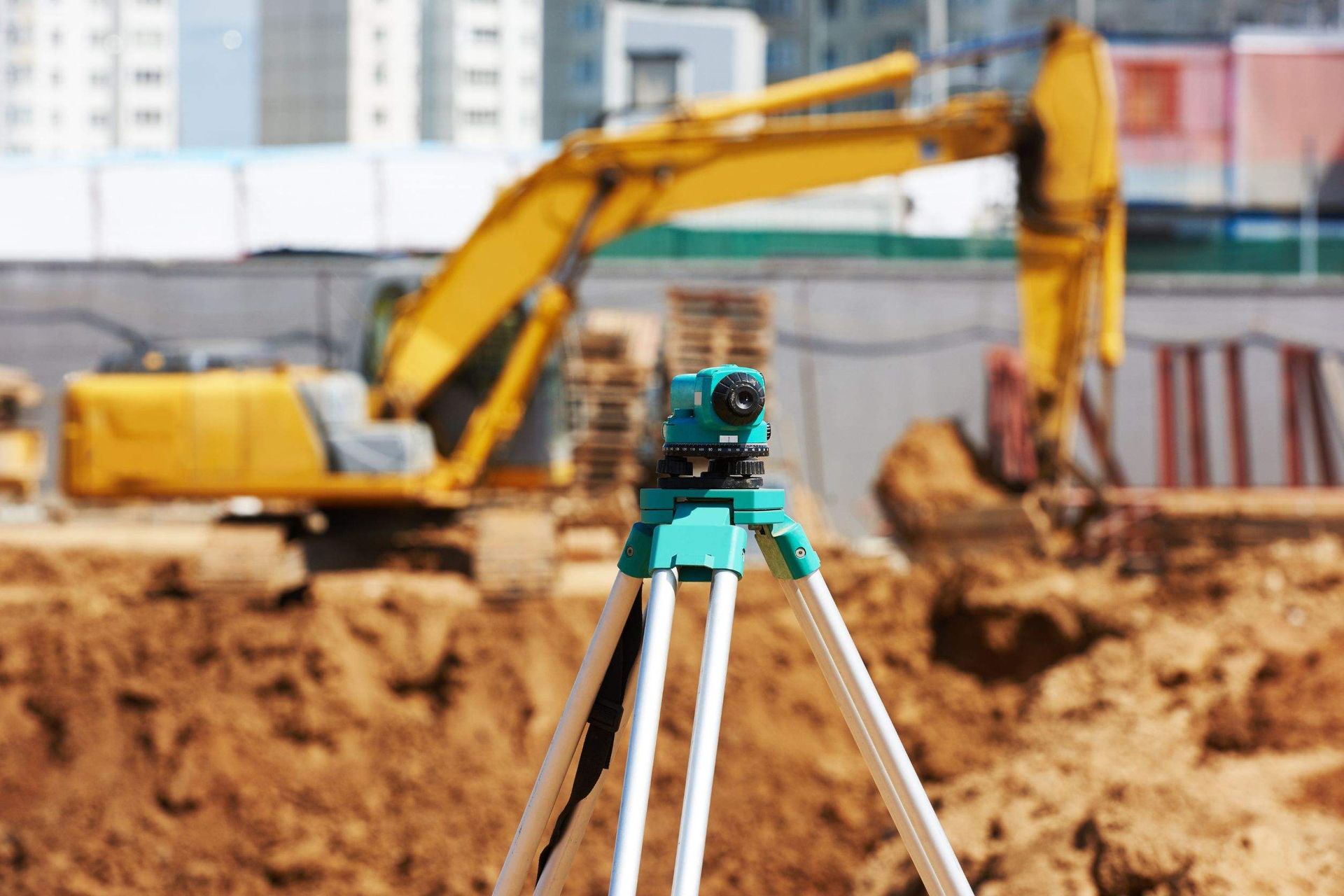 Una obra en construcción con una excavadora amarilla al fondo y un trípode en primer plano.