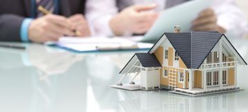 Una pequeña maqueta de una casa sobre una mesa con una persona borrosa escribiendo al fondo.