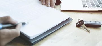 Manos escribiendo en un cuaderno junto a unas gafas y un bolígrafo sobre un escritorio.