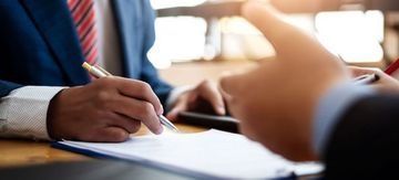 Una persona vestida con un traje azul firma un documento en un escritorio durante una reunión de negocios.