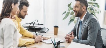 Manos escribiendo en un cuaderno junto a unas gafas y un bolígrafo sobre un escritorio.