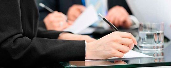 Primer plano de una persona escribiendo en un cuaderno en una mesa de reuniones, con un vaso de agua cerca.