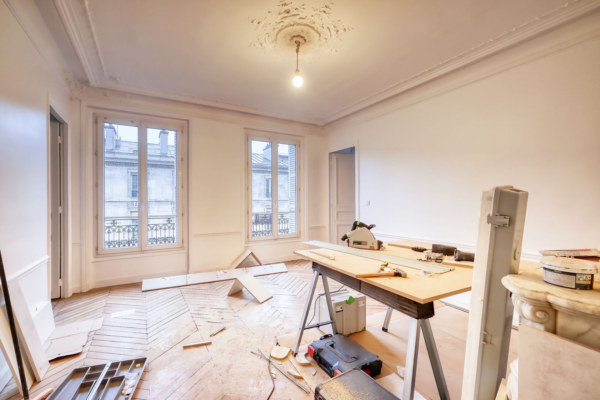 Pièce lumineuse en cours de rénovation, avec outils, bâches de protection et grandes fenêtres.