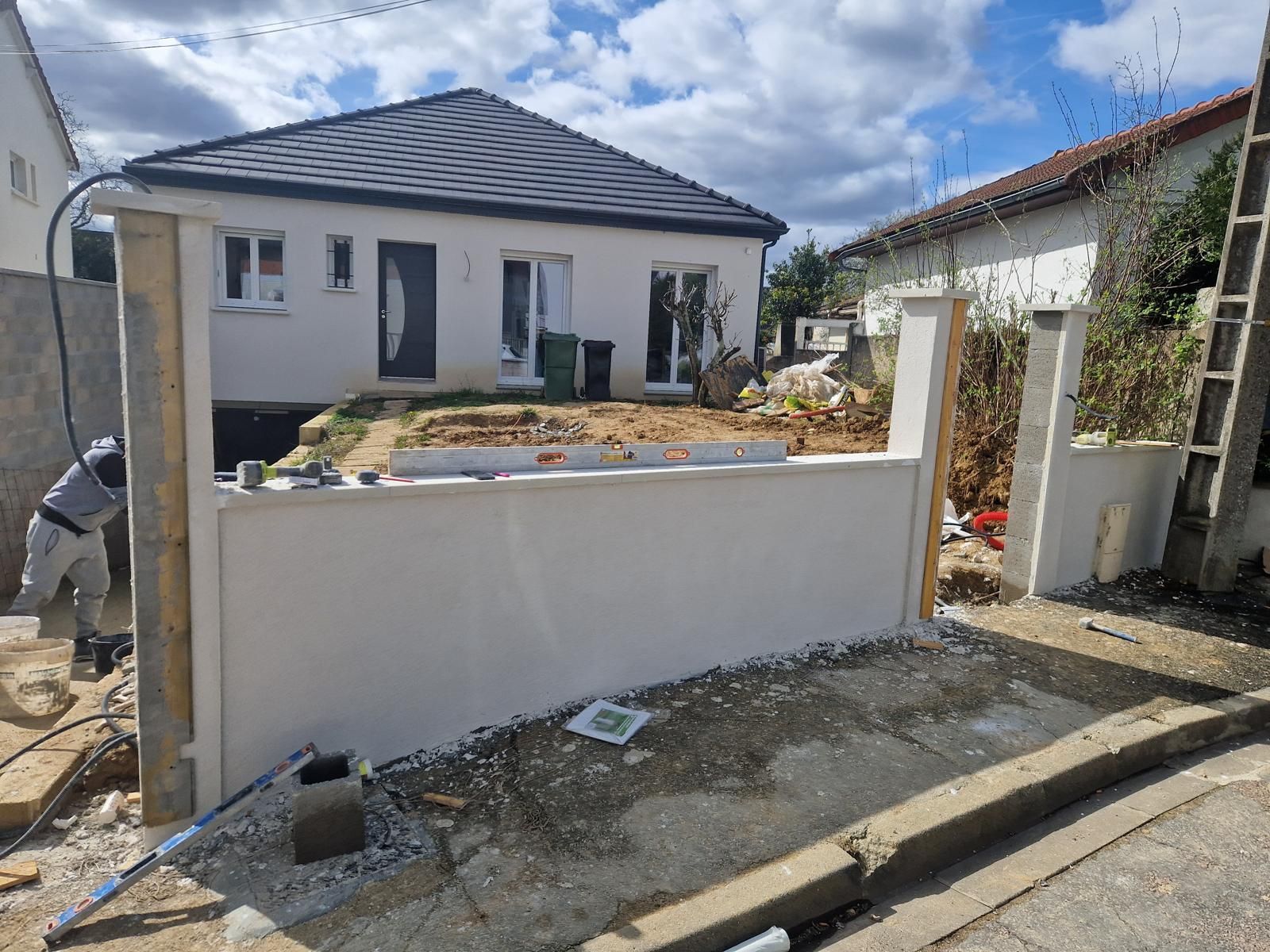Cour avant en travaux avec une maison blanche, un muret et des piliers de portail.