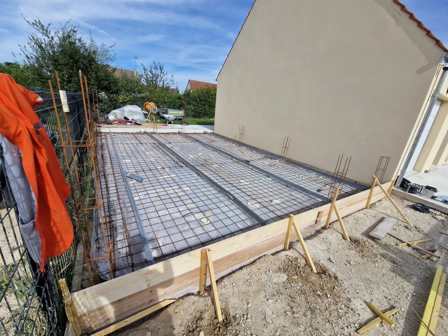 Dalle de fondation en béton en construction à côté d'une maison beige, avec treillis d'armature et coffrage en bois.