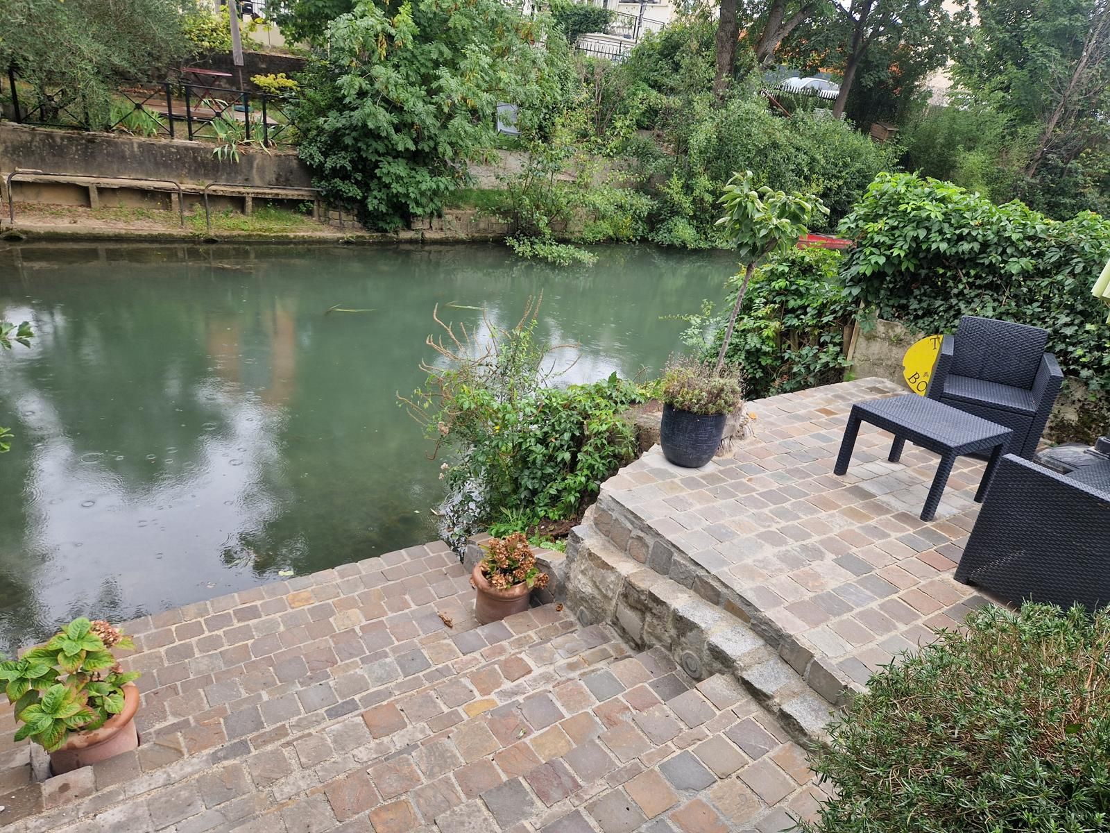 Terrasse en briques dans un jardin luxuriant au bord de la rivière, avec des chaises et une table.