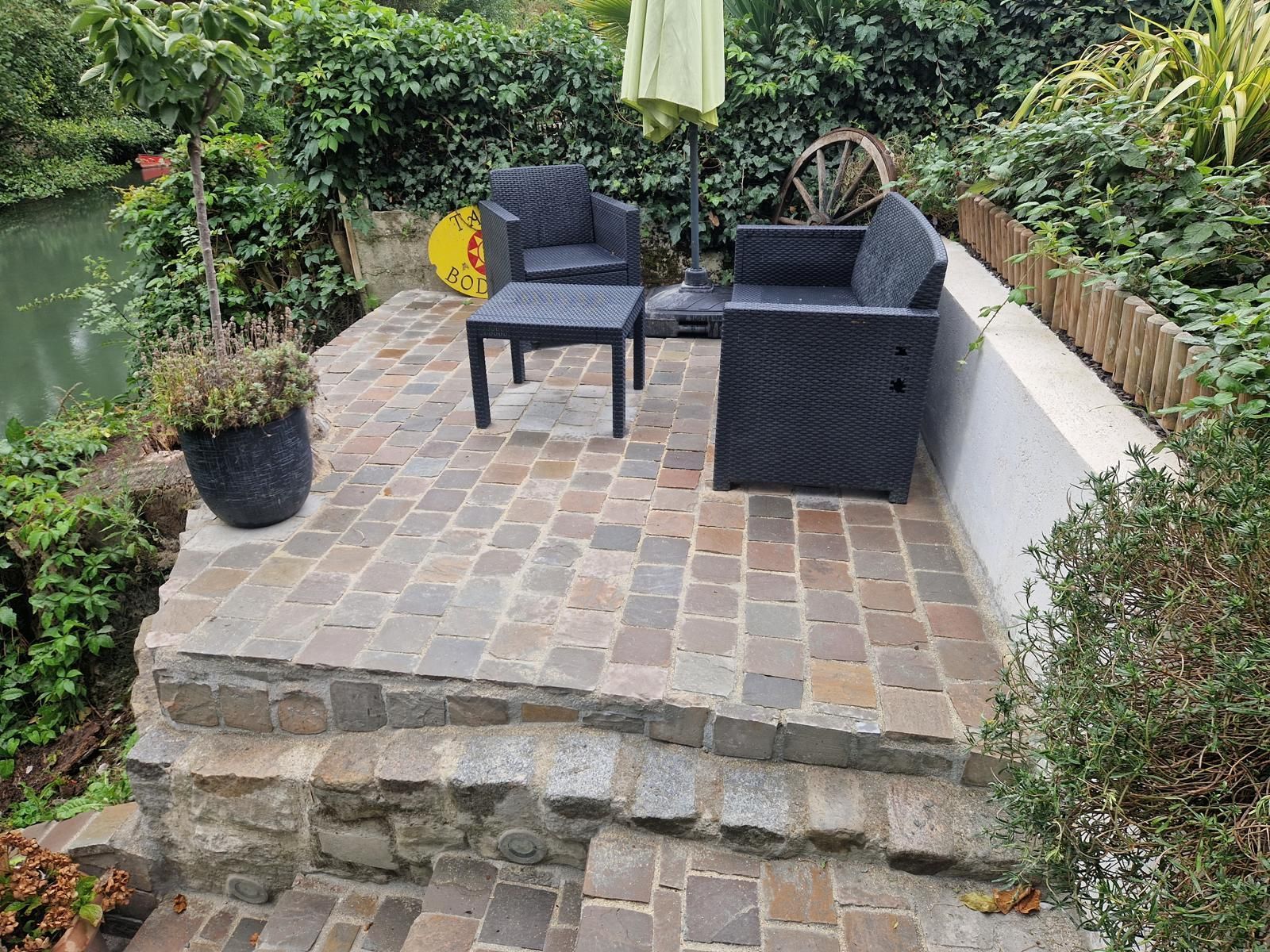 Petite terrasse pavée, chaises de jardin noires, parasol et bordure de jardin luxuriante derrière un muret en pierres.