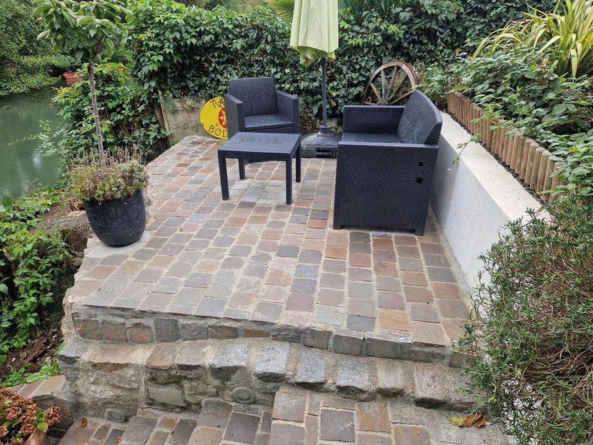Patio avec chaises et table en osier noir sur une terrasse en pavés, entouré d'une végétation luxuriante.