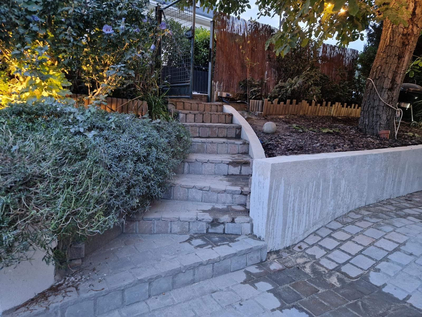 Des marches en pierre, longeant un mur blanc incurvé, mènent à un chemin de jardin bordé d'arbustes et d'arbres.