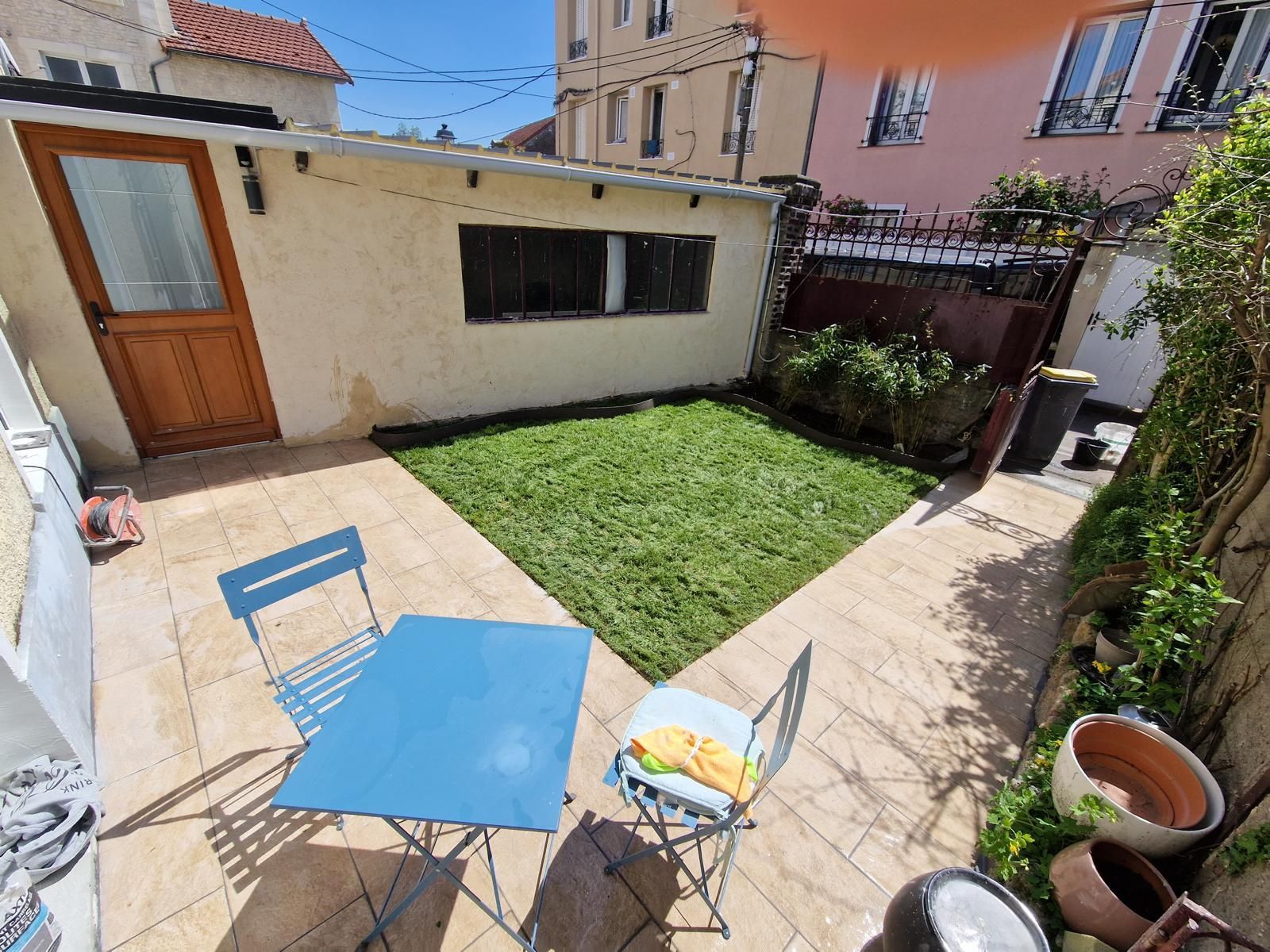 Petite cour avec une table et des chaises bleues, un carré de gazon, une terrasse et des plantes en pot à côté d'une maison.