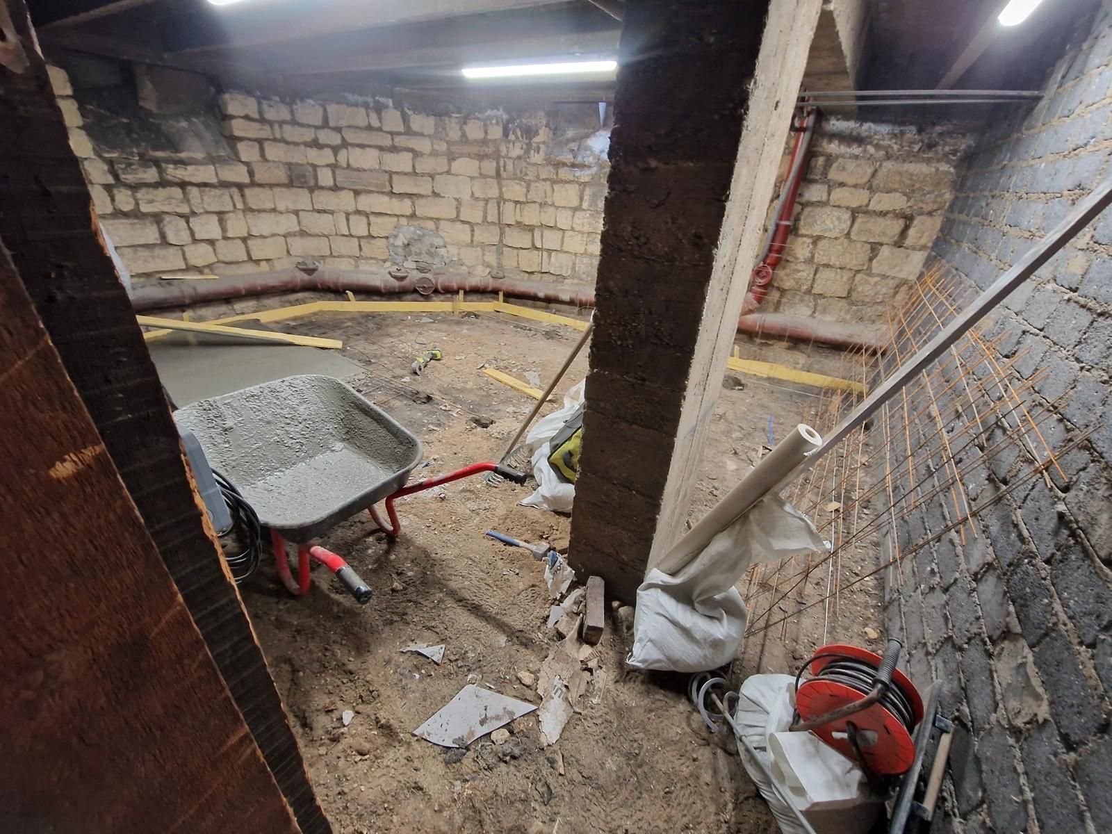 Atelier au sous-sol avec murs en briques apparentes, sol en terre battue, outils et matériaux de construction.