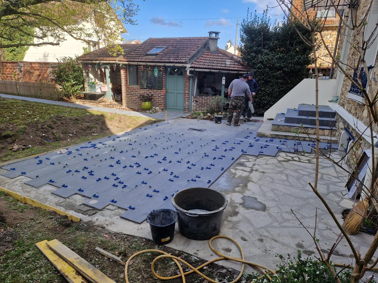 Une terrasse de jardin est aménagée avec des dalles grises à côté d'une petite dépendance en briques et d'un escalier.