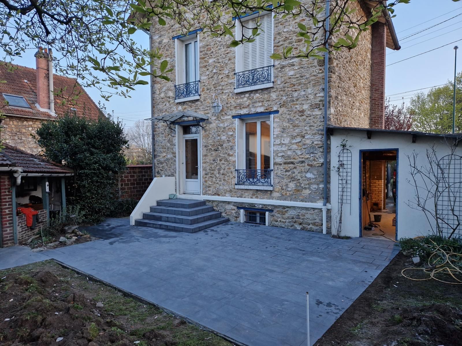 Maison en pierres avec escalier et une extension latérale blanche, vue depuis une cour en dalles.