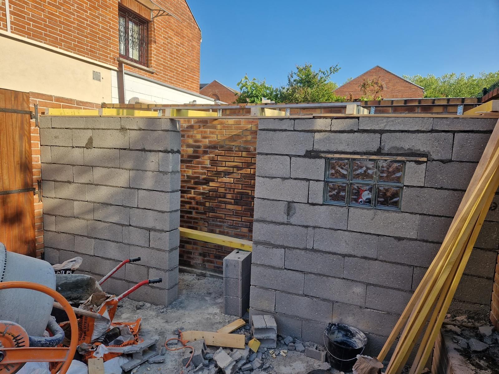 Mur de briques partiellement construit dans une cour arrière, avec des outils et des matériaux de construction.
