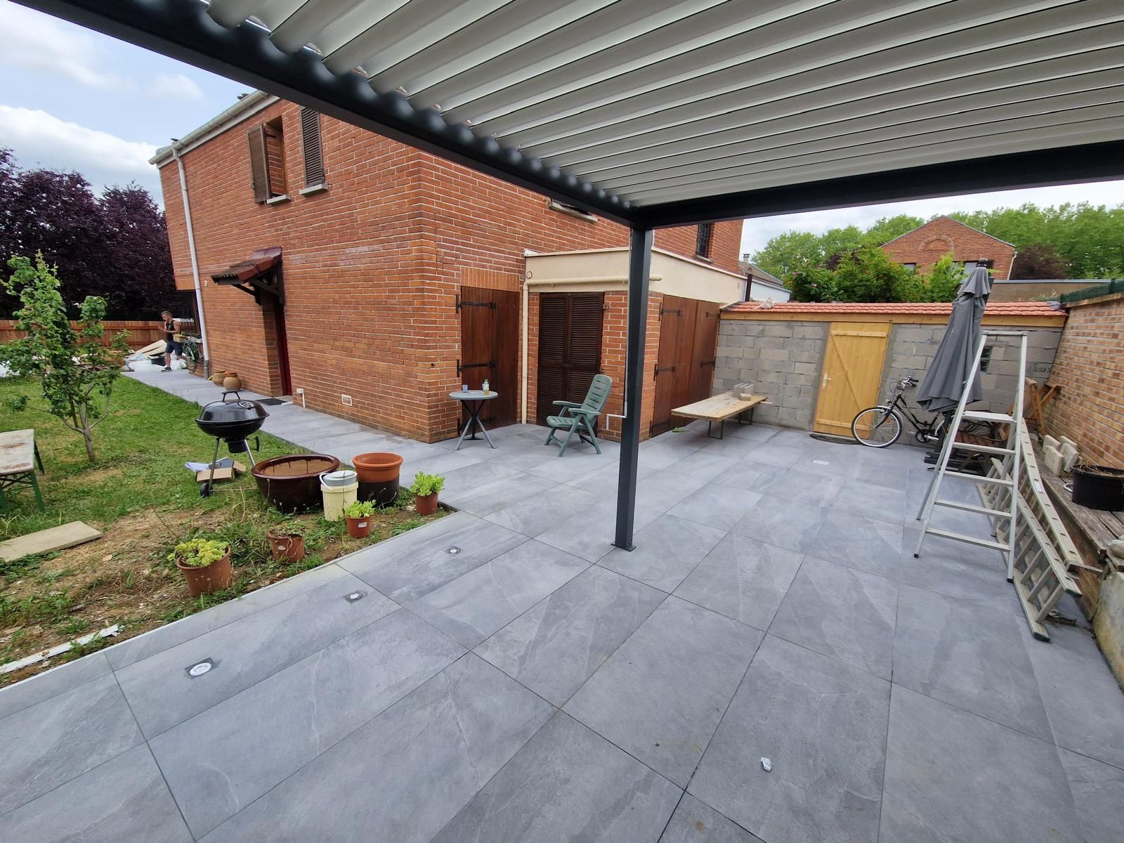 Terrasse couverte avec sol carrelé gris, jouxtant une maison en briques.