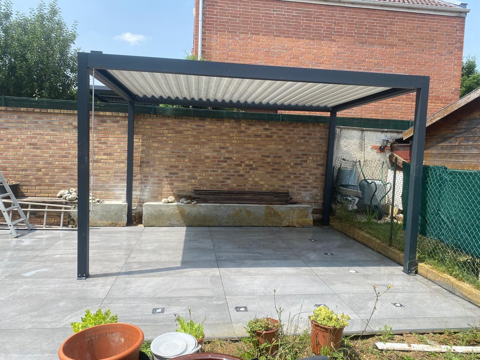 Terrasse arrière avec une pergola en métal noir au-dessus de dalles grises.