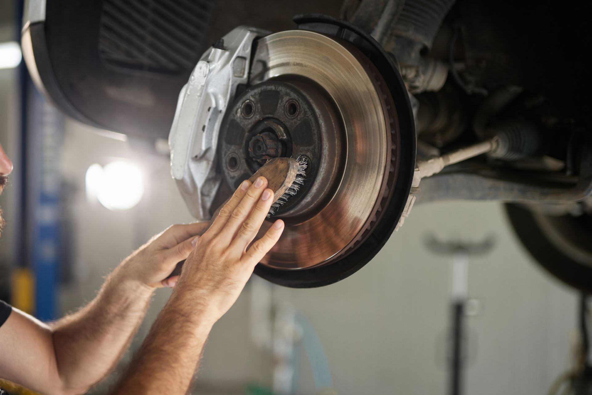 Un mécanicien inspecte un disque de frein. Ses mains touchent le rotor, l'étrier est blanc, la voiture est sur un pont élévateur, dans un atelier.