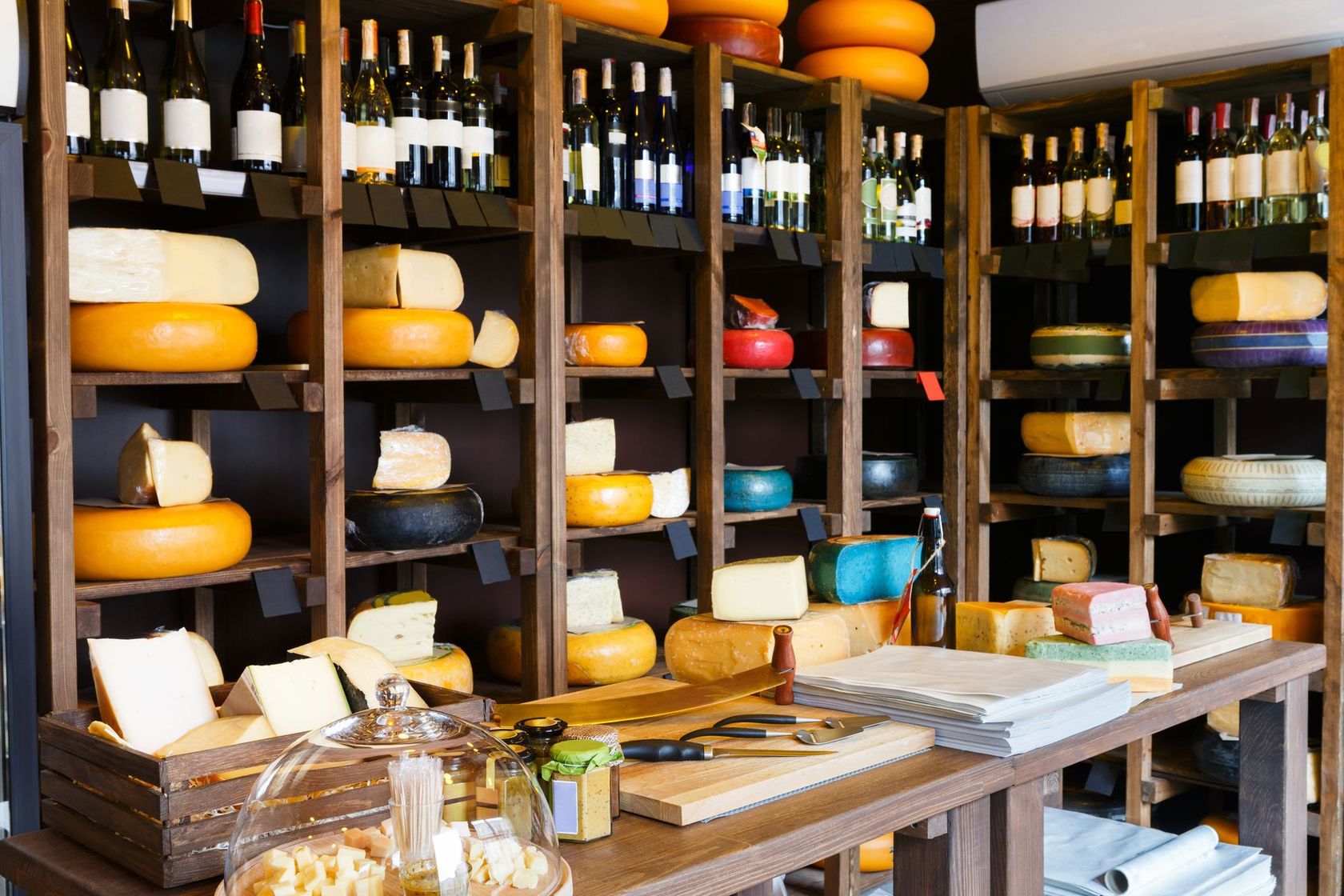 Des étagères regorgent de meules de fromage et de bouteilles derrière un comptoir d'exposition.